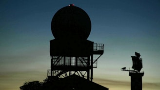 Silhouette of a radar tower with a large circular antenna against a dusk sky. The structure appears complex with metal framework and platforms. In the background, another smaller radar or communication device is visible on the right.