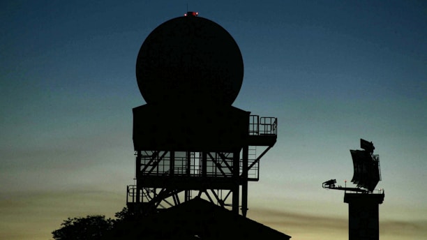 Silhouette of a radar tower with a large circular antenna against a dusk sky. The structure appears complex with metal framework and platforms. In the background, another smaller radar or communication device is visible on the right.