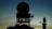 Silhouette of a radar tower with a large circular antenna against a dusk sky. The structure appears complex with metal framework and platforms. In the background, another smaller radar or communication device is visible on the right.