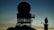 Silhouette of a radar tower with a large circular antenna against a dusk sky. The structure appears complex with metal framework and platforms. In the background, another smaller radar or communication device is visible on the right.