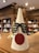 A paper cone decorated as a humorous reindeer, complete with black shutter shades and a large red circle on the front, resembling a nose. The structure has brown antlers attached to the top. In the background, there are bookshelves filled with books in a well-lit room.