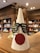 A paper cone decorated as a humorous reindeer, complete with black shutter shades and a large red circle on the front, resembling a nose. The structure has brown antlers attached to the top. In the background, there are bookshelves filled with books in a well-lit room.