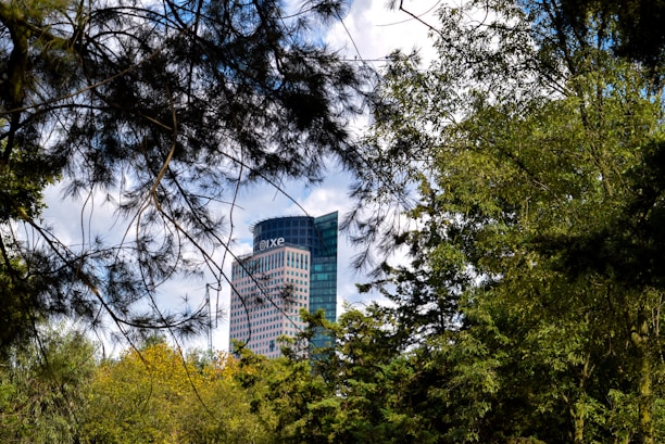 A modern skyscraper with a rounded glass design is partially obscured by lush green trees and foliage. The building bears the name 'Río Tiber 100'. The sky above is partly cloudy, adding a soft contrast to the vibrant greenery.