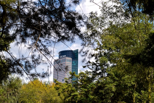 A modern skyscraper with a rounded glass design is partially obscured by lush green trees and foliage. The building bears the name 'R&iacute;o Tiber 100'. The sky above is partly cloudy, adding a soft contrast to the vibrant greenery.