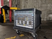 A large networking device, likely a server or network switch, is positioned on a small wheeled platform in an industrial setting with concrete walls and a visible yellow-painted structural support. The equipment features a series of component slots and ports arranged in rows on its front panel.