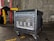 A large networking device, likely a server or network switch, is positioned on a small wheeled platform in an industrial setting with concrete walls and a visible yellow-painted structural support. The equipment features a series of component slots and ports arranged in rows on its front panel.