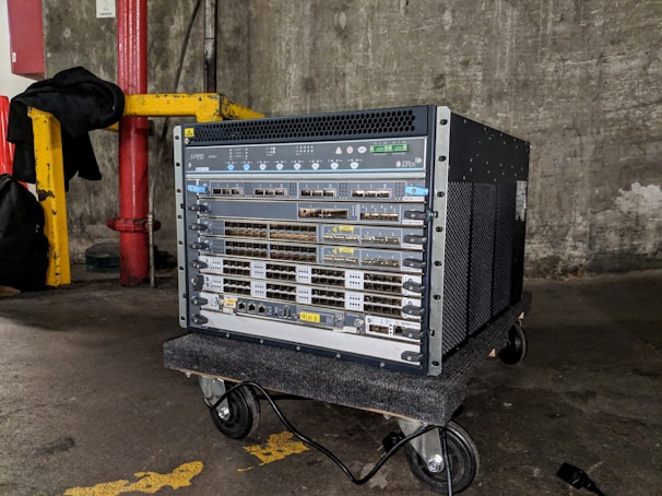 A large networking device, likely a server or network switch, is positioned on a small wheeled platform in an industrial setting with concrete walls and a visible yellow-painted structural support. The equipment features a series of component slots and ports arranged in rows on its front panel.