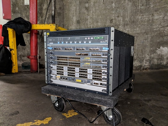 A large networking device, likely a server or network switch, is positioned on a small wheeled platform in an industrial setting with concrete walls and a visible yellow-painted structural support. The equipment features a series of component slots and ports arranged in rows on its front panel.