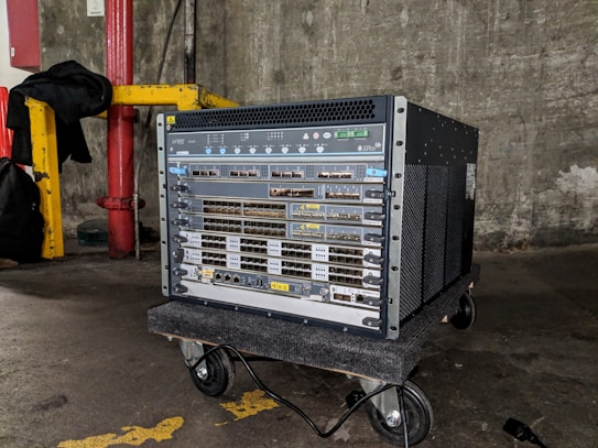 A large networking device, likely a server or network switch, is positioned on a small wheeled platform in an industrial setting with concrete walls and a visible yellow-painted structural support. The equipment features a series of component slots and ports arranged in rows on its front panel.