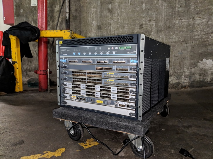 A large networking device, likely a server or network switch, is positioned on a small wheeled platform in an industrial setting with concrete walls and a visible yellow-painted structural support. The equipment features a series of component slots and ports arranged in rows on its front panel.