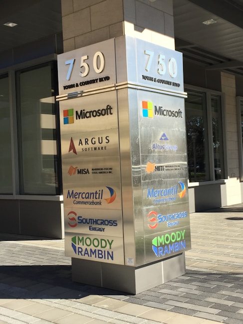 A tall, rectangular signpost displays the address 750 Town & Country Blvd and lists several company logos including Microsoft, Argus Software, Altus Group, MISA, Mercantil Commercebank, Southcross Energy, and Moody Rambin. The signpost is set against a modern office building with large windows and tiling on the ground.