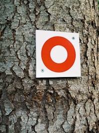 A textured tree trunk with a square white sign attached. The sign features a prominent red circle in the center, secured with two screws at the corners.