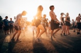 A group of people enjoying an outdoor music festival, some dancing and others standing. The sun is setting, casting a warm glow over the scene, and creating an energetic and carefree atmosphere.