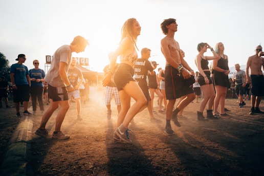 A group of people enjoying an outdoor music festival, some dancing and others standing. The sun is setting, casting a warm glow over the scene, and creating an energetic and carefree atmosphere.