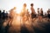 A group of people enjoying an outdoor music festival, some dancing and others standing. The sun is setting, casting a warm glow over the scene, and creating an energetic and carefree atmosphere.
