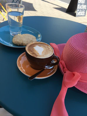 A steaming cup of cappuccino on a pastel pink table next to beauty products.