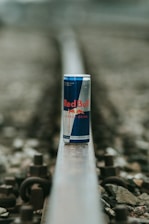 A can of Red Bull energy drink is placed on a railway track. The focus is on the can, while the surrounding environment, including the rail and gravel, is blurred. The can features the distinctive Red Bull branding with blue and silver colors.
