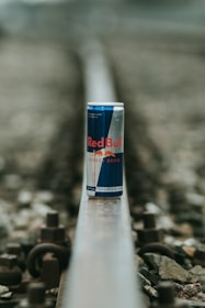 A can of Red Bull energy drink is placed on a railway track. The focus is on the can, while the surrounding environment, including the rail and gravel, is blurred. The can features the distinctive Red Bull branding with blue and silver colors.