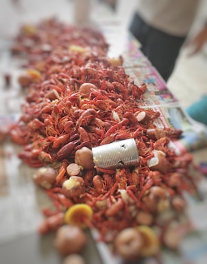 A large spread of boiled crawfish, potatoes, and other vegetables on a newspaper-covered table. In the center lies an empty tin can. The setup suggests a traditional crawfish boil with a casual and communal atmosphere.