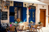 An outdoor cafe scene with blue doors and windows enhanced by colorful flower pots. The seating area includes wicker chairs and a glass table. A chalkboard menu displays breakfast items and specials. A quaint, Mediterranean atmosphere is suggested by the decorative elements and the rustic wall texture.