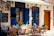 An outdoor cafe scene with blue doors and windows enhanced by colorful flower pots. The seating area includes wicker chairs and a glass table. A chalkboard menu displays breakfast items and specials. A quaint, Mediterranean atmosphere is suggested by the decorative elements and the rustic wall texture.