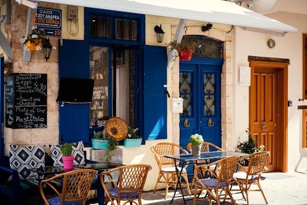 An outdoor cafe scene with blue doors and windows enhanced by colorful flower pots. The seating area includes wicker chairs and a glass table. A chalkboard menu displays breakfast items and specials. A quaint, Mediterranean atmosphere is suggested by the decorative elements and the rustic wall texture.