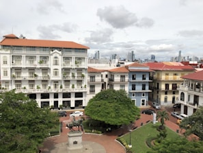 A picturesque urban scene featuring a well-preserved historic district with colorful colonial-style buildings. The architecture includes ornate details and balconies, surrounded by lush green trees and a pedestrian plaza. A statue of a person on horseback is prominently placed in the center of the plaza. The skyline of a modern city is visible in the background under a cloudy sky.