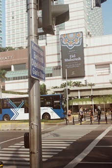 A city street scene with a large 'Eid Mubarak' billboard on a building, featuring a geometric design and text in multiple languages. Below the billboard, there is a traffic light and a blue public bus near a pedestrian crossing, where a few people are standing. A sign in a different language is attached to a pole.