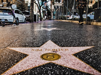 A perspective view of a star on the Hollywood Walk of Fame with the name Kim Hunter. The image shows a sidewalk with other stars lining the path. There are cars parked along the street, a scooter leaning against a car, and a person walking by. The background features buildings, fencing, and palm trees in an urban setting.