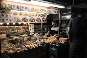 A street food vendor is standing at a stall filled with various fried snacks. The vendor is preparing or arranging food items on orange plates. The stall has a bright, illuminated menu displaying images of the snacks being sold, with some descriptions written in another language. The atmosphere is dimly lit, suggesting it is evening.