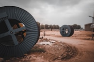 Large industrial cable reels are lying on a dirt road in an open area. The sky is overcast, giving a gloomy atmosphere. A solitary person is visible in the distance, walking on the muddy path. Surrounding the area are various trees and industrial structures.