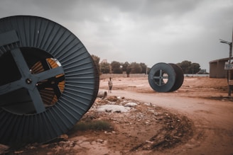 Large industrial cable reels are lying on a dirt road in an open area. The sky is overcast, giving a gloomy atmosphere. A solitary person is visible in the distance, walking on the muddy path. Surrounding the area are various trees and industrial structures.