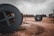 Large industrial cable reels are lying on a dirt road in an open area. The sky is overcast, giving a gloomy atmosphere. A solitary person is visible in the distance, walking on the muddy path. Surrounding the area are various trees and industrial structures.
