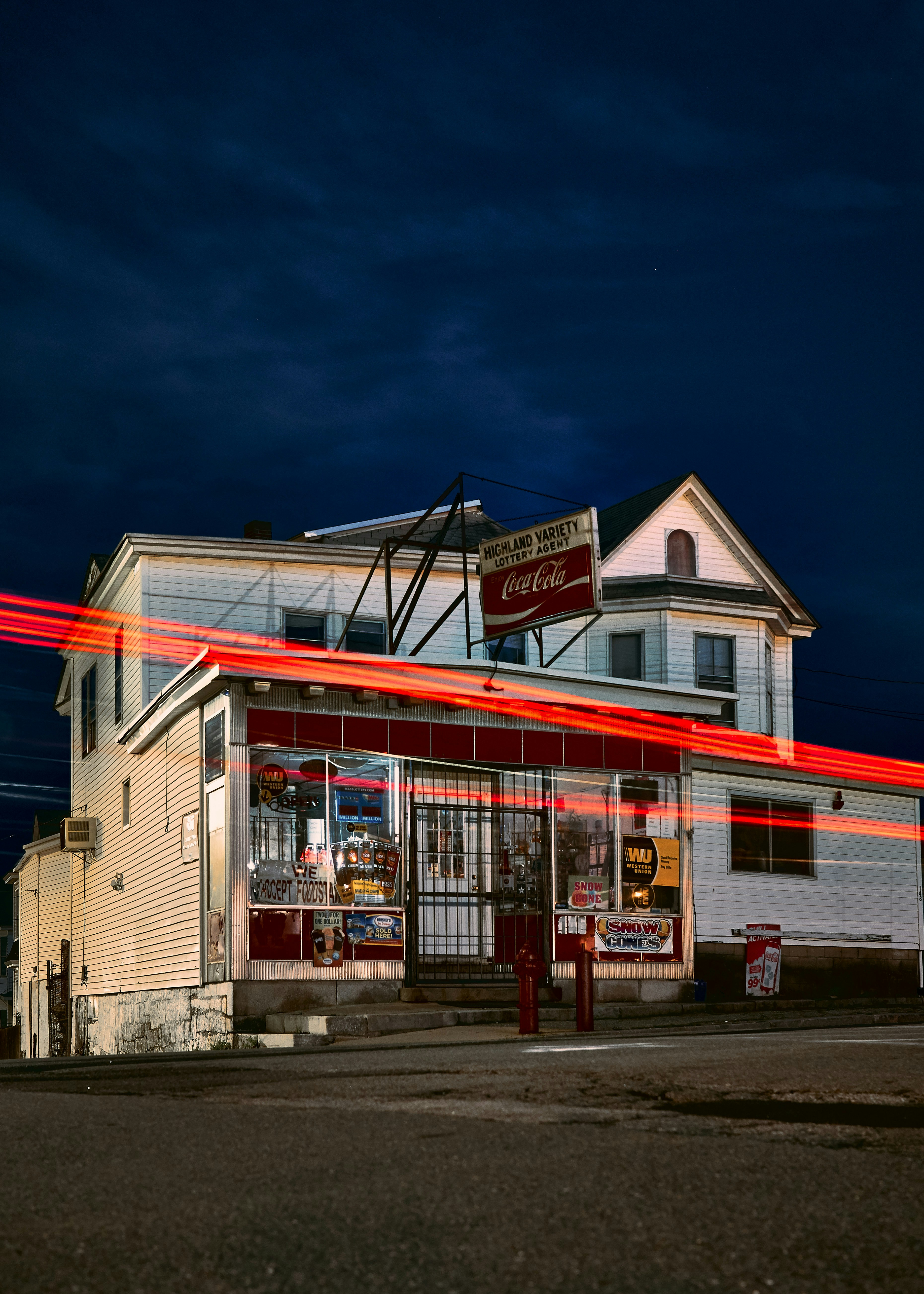 Retro two-story corner store at night, its storefront awash in warm interior light with a Coca-Cola sign visible. A vivid red light trail crosses the façade, creating motion against the dark evening sky.