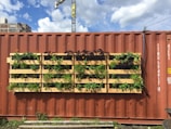 A large red shipping container with a vertical garden made of wooden pallets attached to its side. The pallets are filled with various green plants and small flowers, thriving under sunlight. Some industrial buildings and a construction crane are visible in the background against a partly cloudy sky.