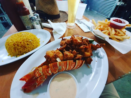 An elegant table setting features a meal consisting of fried seafood, possibly lobster, with a colorful shell on a white plate accompanied by a small bowl of dipping sauce. A mound of yellow rice is served on a separate plate and a basket of golden fries with ketchup is visible. A tall glass of beer or light beverage stands in the background next to condiments like hot sauce and salt. A straw hat rests on the table, adding a casual touch to the dining experience.