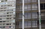 A close-up of two contrasting building facades, with the left side showing a residential apartment complex with a series of balconies and windows. The right side features an office-like glass facade with a Brazilian flag attached and security cameras mounted.