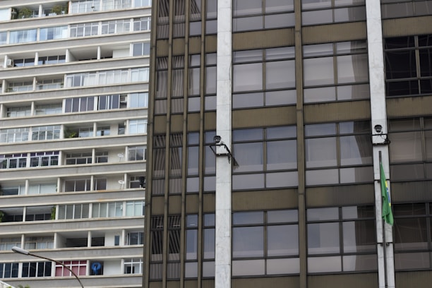 A close-up of two contrasting building facades, with the left side showing a residential apartment complex with a series of balconies and windows. The right side features an office-like glass facade with a Brazilian flag attached and security cameras mounted.