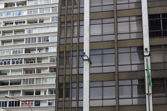 A close-up of two contrasting building facades, with the left side showing a residential apartment complex with a series of balconies and windows. The right side features an office-like glass facade with a Brazilian flag attached and security cameras mounted.