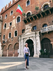 A person stands in front of a historic brick building with arched windows and decorative stonework. An Italian flag is attached to the building, fluttering in the wind. The individual is wearing sunglasses, a striped shirt, and a blue skirt, and carries a small bag. Potted plants are visible on a balcony above.