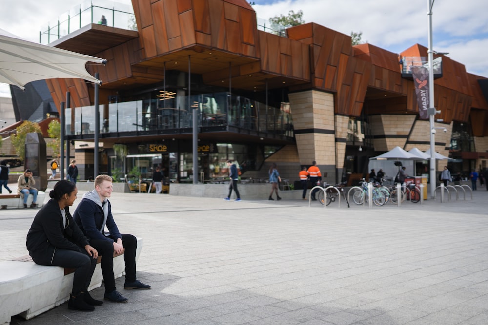 man and woman sitting near water fountain beside people walking near building during daytime