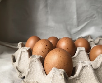 Several brown eggs are placed in a grey, textured egg carton. The background is soft and neutral, with gentle lighting accentuating the smooth surface of the eggs.