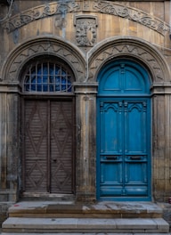 Two distinct doors are set side by side, each framed by ornate arches. The left door is wooden with a geometric diamond pattern, while the right door is vibrant blue with intricate paneling. Above the doors, a relief sculpture and decorative carvings enhance the aged facade.