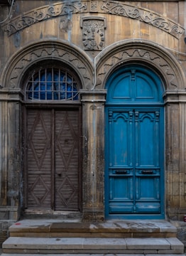 Two distinct doors are set side by side, each framed by ornate arches. The left door is wooden with a geometric diamond pattern, while the right door is vibrant blue with intricate paneling. Above the doors, a relief sculpture and decorative carvings enhance the aged facade.