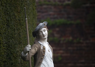 A statue of a historical figure wearing 18th-century attire, including a tricorn hat and holding a long staff or spear. The statue is positioned against a backdrop of a brick wall with creeping ivy.