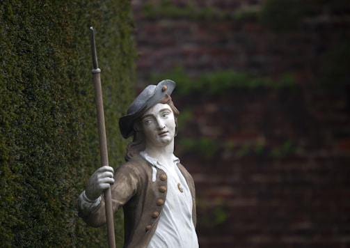 A statue of a historical figure wearing 18th-century attire, including a tricorn hat and holding a long staff or spear. The statue is positioned against a backdrop of a brick wall with creeping ivy.