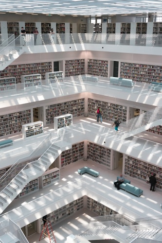 A modern, multi-level library with white walls and shelves filled with books. Sunlight streams through the ceiling, casting patterns on the floors and railings. Several people are browsing bookshelves or walking along the corridors, while others are seated on light blue couches placed around the library.