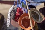 A collection of various hats is displayed, hanging from blue plastic clips attached to a pole. The hats include different styles and colors, such as a striped one, a floral red one, and straw hats. Sunlight highlights the textures of the hats and casts shadows, creating contrasting colors and patterns.