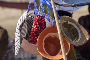 A collection of various hats is displayed, hanging from blue plastic clips attached to a pole. The hats include different styles and colors, such as a striped one, a floral red one, and straw hats. Sunlight highlights the textures of the hats and casts shadows, creating contrasting colors and patterns.