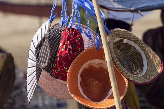 A collection of various hats is displayed, hanging from blue plastic clips attached to a pole. The hats include different styles and colors, such as a striped one, a floral red one, and straw hats. Sunlight highlights the textures of the hats and casts shadows, creating contrasting colors and patterns.